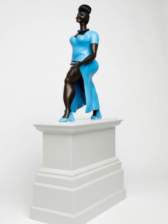 Figure or a black woman, in a blue dress and blue shoes, walking forward, on a light grey plinth, back view
