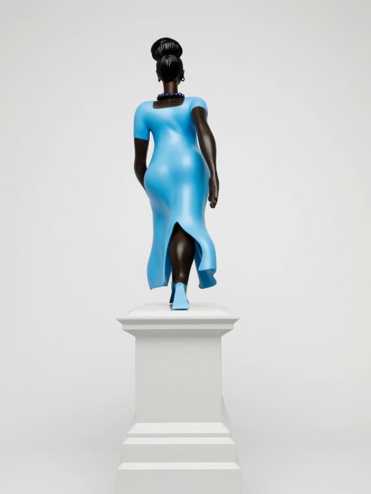 Figure or a black woman, in a blue dress and blue shoes, walking forward, on a light grey plinth, side angled view