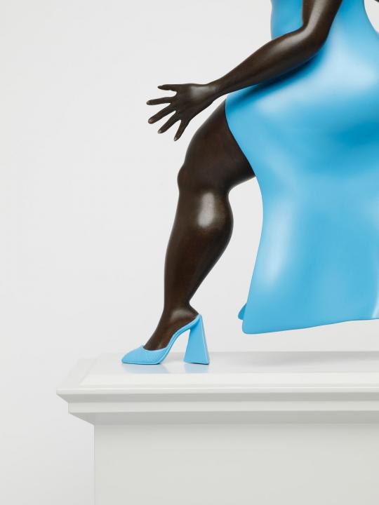 Figure or a black woman, in a blue dress and blue shoes, close up view of her arm and leg walking forward, on a light grey plinth.