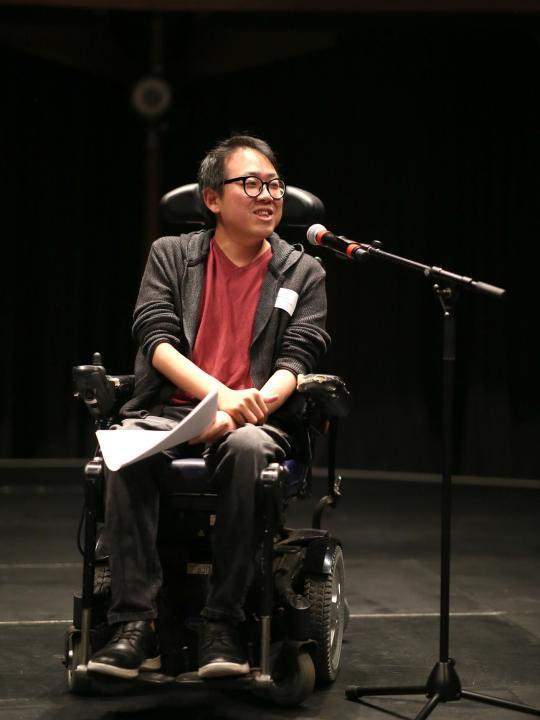 James Lee speaks into a standing microphone, smiling as he addresses the audience. He is wearing black jeans, a red top and black jumper, lit up by warm stage lighting in his mobility chair against a dark background.