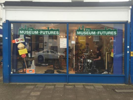 The Museum of Futures in Surbiton shop window displaing art and a musician playing the drums.