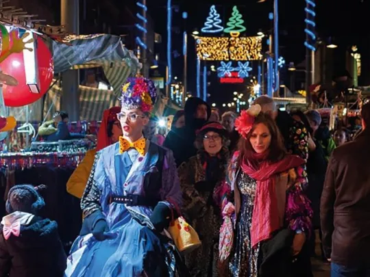 Walthamstow Market at night. Crowds of people shopand socialise. In the front of the picture someone dressed in a bright headress and blue dress.