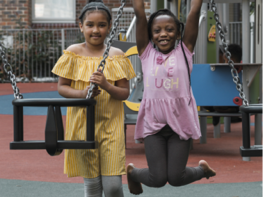 Two girls smiling at camera as they play on the swings in a playground