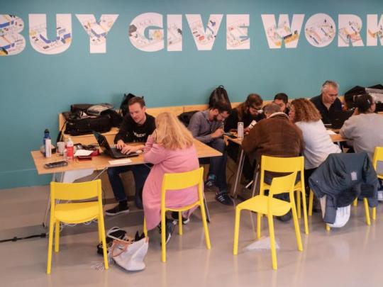 A room at International House in Brixton. A blue wall with large letters saying BUY GIVE WORK as young people sit at tables working together.