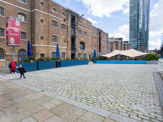 A cobbled public square next to a 4 storey tan-coloured brick block building next to a cobbled public square in London Docklands. In the distance is a glass tower block and cafe area.