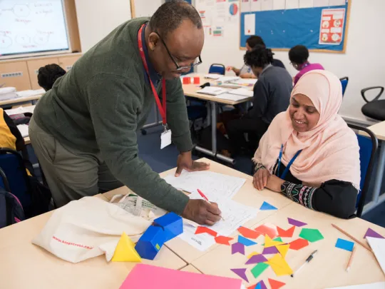 A tutor helps a young learner at her classroom desk, which is filled with colourful shapes and papers.