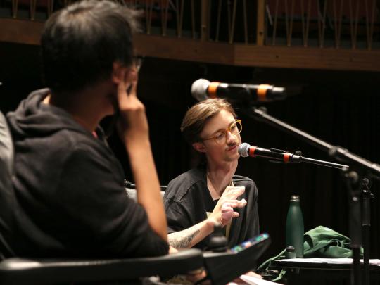 Two men engaged in conversation on stage, the camera looks over one mans shoulder to the other man who's speaking into a mic.