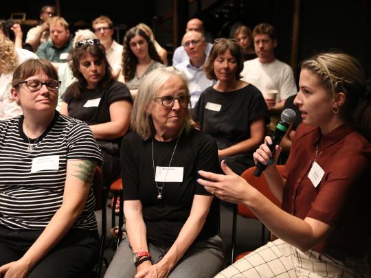 Audience member sits holding a mic, addressing the rest of the audience to her right.