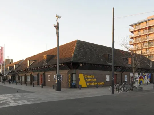 The exterior of the Albany building as viewed from Douglas Way - it is a low level brick building with pink flags above the entrance and yellow text on the wall that reads Theatre. Cafe Bar. Creative Space.