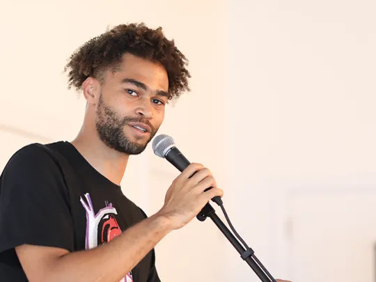 A performer with curly hair holds a microphone and looks into the camera. 