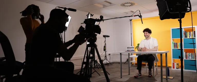 Behind the scenes of filming; a camera and its handler filming a presenter sat at a desk with lighting and mic visible.