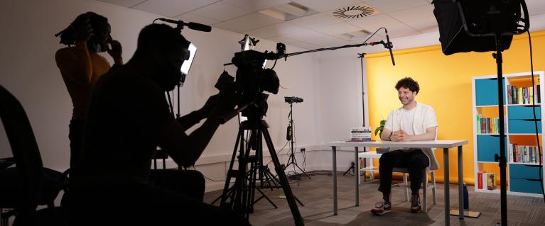 Behind the scenes of filming; a camera and its handler filming a presenter sat at a desk with lighting and mic visible.