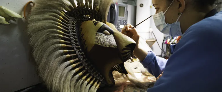 A person with a paint brush in their hand, brushing a lion head piece from Disney's The Lion King theatre
