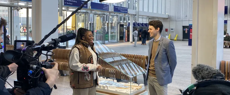 Behind the scenes of an interview. A person handling a camera and another holding lighting above two people; an interviewer and interviewee.