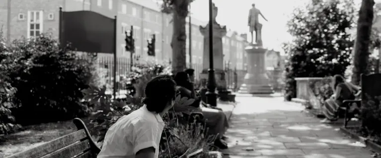 A woman sat in a park in Bow, with headphones in, listening to audio story about the Gladstone statue which is in the background