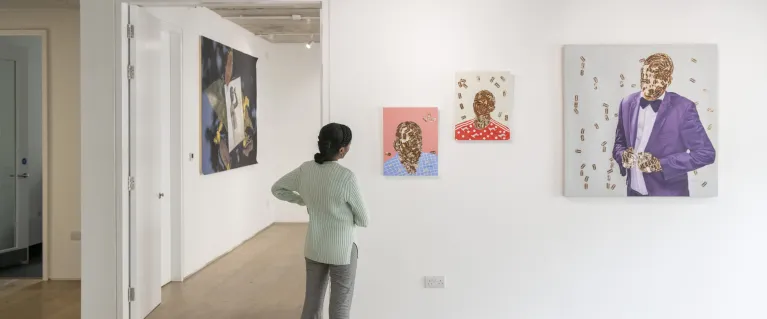 A person gently cocks their head to admire three abstract portrait paintings Black displayed on an art gallery wall.
