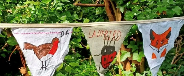 Children's drawings of a robin, ladybird and fox on bunting strung in a woodland.