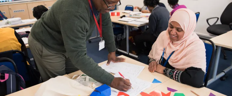 A tutor helps a young learner at her classroom desk, which is filled with colourful shapes and papers.