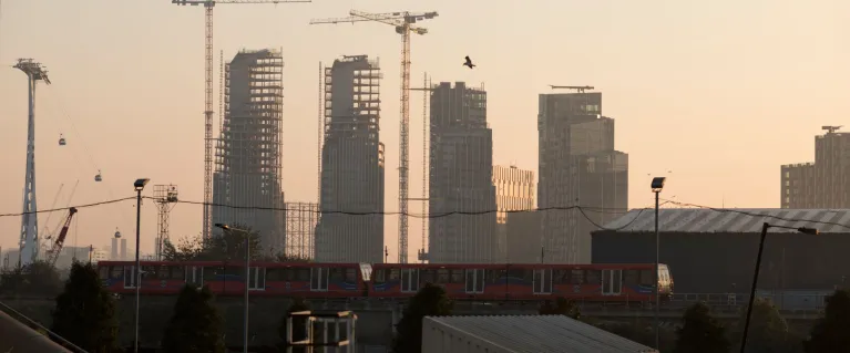 Construction site of 4 tower blocks at dusk. DLR train passing in front, cable car overhead lines to the left.