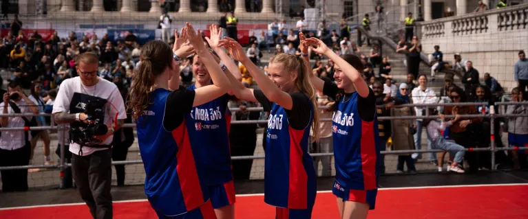 Four female players in blue and red Junior NBA 3v3 uniforms celebrate with high-fives on a basketball court in Trafalgar Square, surrounded by spectators.