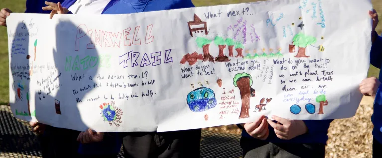 A group of children hold up a banner for “Pinkwell Trail”. The banner has children’s drawings of trees and text explaining what they want from their nature trail.