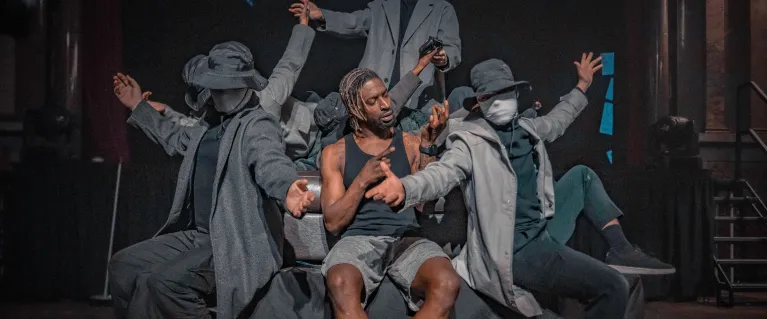 Sim Campbell sits centre stage on a sofa. He is a black man with dyed dreadlocks, wearing a black vest, grey shorts and white socks. He's surrounded by mysterious figures wearing trench coats, masks and hats. They frame him as they pass objects to eachother, behind and around him. But Sim is looking intently at his own hands, which are held up in front of him as he uses sign language.