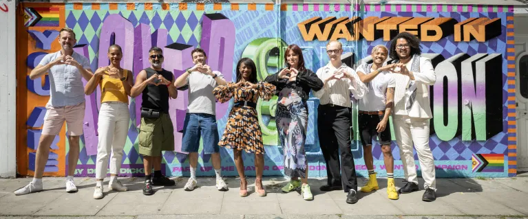 Group of people standing in front of a colourful Loved and Wanted mural, holding their hands up to form love hearts.