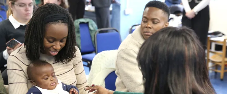 Three people are sitting down on chairs around each other, one of which has a child in her lap