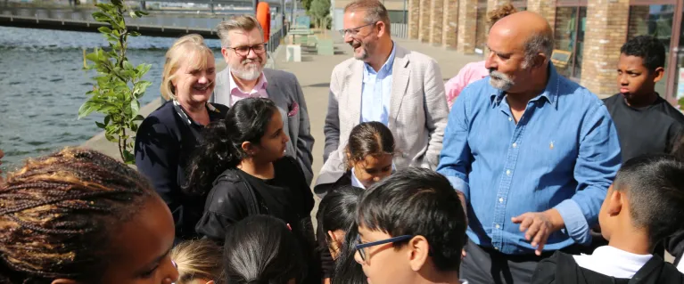 A group of 12 people, most of which are children, stand by the river and have a building behind them
