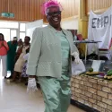 An older black woman walks proudly to camera, smiling, wearing a silk green dress, jacket, with lace gloves, and pink hair fascinator