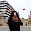 A young white woman wearing a large black hooded jacket, she looks in the distance, a backdrop of buildings and urban bridges and structures