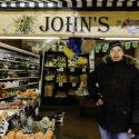 A middle aged white man wearing a black coat and woolly hat stands in a grocer’s shop amongst fruit and veg with a sign saying ‘John’s’ above him.