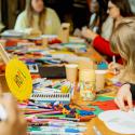 Close up of a busy table top with coloured pens and arts and crafts tools. There are people around the table, including a child drawing, and a sign at the forefront reads ‘hope’