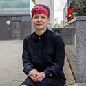 Zoë Garbett sits on a stone bench outside in London, staring straight at the camera ahead without smiling. Her legs are crossed and her hands are in her lap.