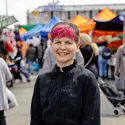Zoë Garbett stands on a sunny day at Ridley Road Market in London. She is smiling straight at the camera wearing a black longsleeve blouse.
