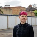 Zoe Garbett stands in front of a block of London residential garages painted white. Zoe is turned to the right of the camera staring in the distance. She is wearing a black blouse.