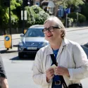 Caroline Russell campaigning for more push buttons on street crossings across London. She is smiling, wearing a white jacket and looking off to the left of the camera engaging with a Londoner. A car is visible in the background. 