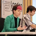 Zoë Garbett AM is seen chairing London's first rent commission in the chamber at City Hall. She is wearing a green blazer, a black shirt, and has her trademark pink fringe.
