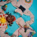 A young person with colourful braids lies on the floor looking straight up at the camera with a wide smile, wearing a hoodie with a sun that says ‘OK’ and holding two other hoodies with colourful writing and beads spread on the floor. It is vibrant and colourful.
