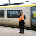 A member of staff, wearing a high-visibility jacket, stands on a station platform as a ThamesLink train rolls past.