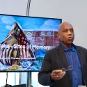 Burt Caesar delivers a talk in front of an illustration of a cowrie shell shaped memorial