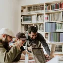 A group of people chat and laugh sat around a table with laptops in a library