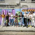 Group of people standing in front of a colourful Loved and Wanted mural, holding their hands up to form love hearts.