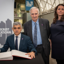 Sadiq Khan seated signing a book of commitment with Karen Pollock stood next to him