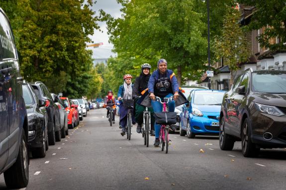 A group of women cycling along rows of parked cars.