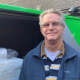 A man wearing a casual jacket and striped shirt, with grey hair and glasses, stands in front of a bright green "Zip" van.  The sliding side door of the van is open to show two cot mattresses stacked inside.  The man is smiling and facing the camera.